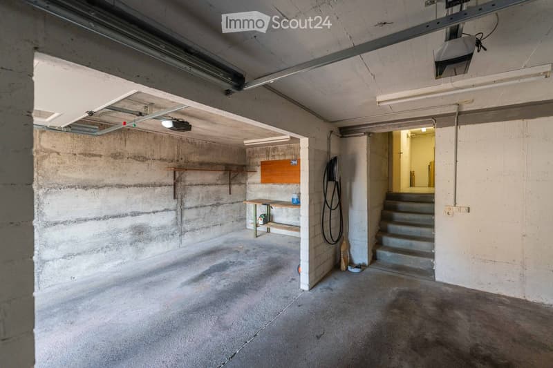 Empty concrete-walled garage with a small wooden workbench, shelves, and a broom. Stairs lead to another area.
