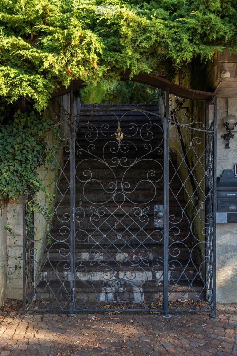 An ornate wrought iron gate with decorative scrollwork and floral motifs leads to a stone staircase, surrounded by lush greenery and ivy.