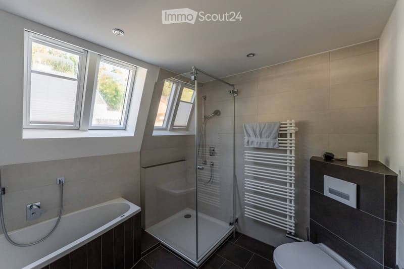 Modern bathroom with a bathtub, toilet, glass-enclosed shower, radiator, and towel on a shelf. The room features large windows and a sleek design.