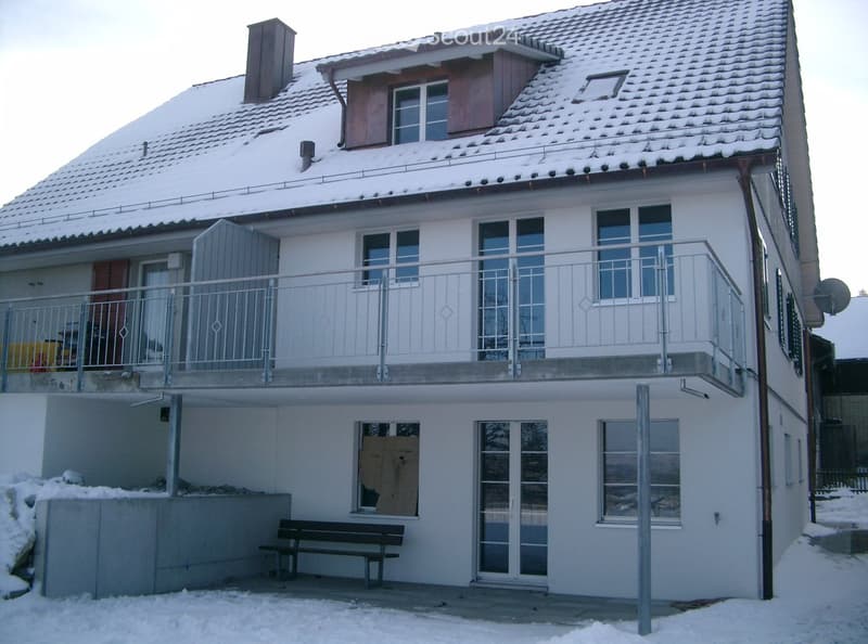 Two-story house with white exterior, snow-covered roof, balcony with metal railing, and a bench near the entrance.