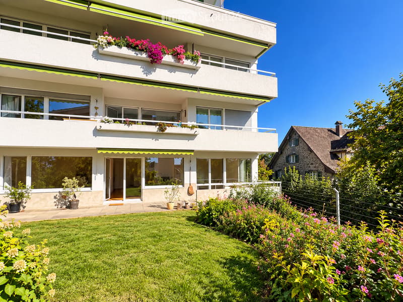 Modern three-story apartment building with balconies, large windows, and a well-maintained garden with green lawn and colorful flowers.