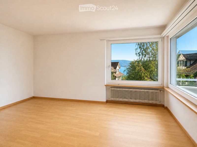 Empty room with light wooden flooring, white walls, large windows showing a scenic view, and a radiator below one window.
