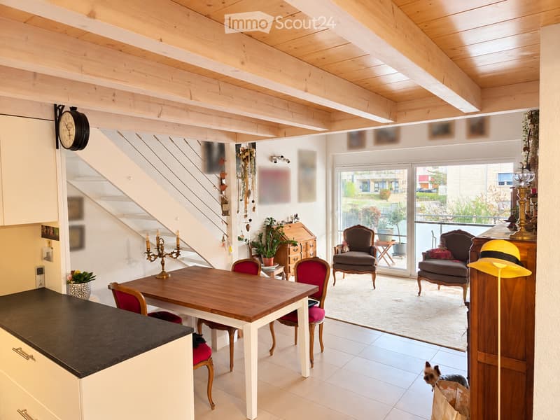 Bright, open-plan living and dining area with exposed wooden beams, a dining table with chairs, a candle holder, a clock, and a small dog visible near the entrance to a balcony.
