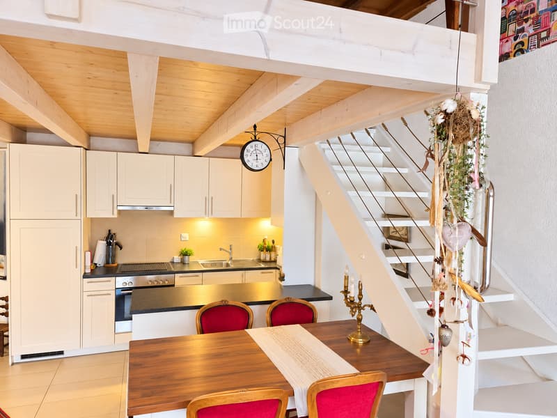 Bright, modern kitchen with white cabinets, stainless steel appliances, and a wooden dining table with red chairs. A staircase leads to an upper level, decorated with a hanging floral arrangement and a candle holder on the steps.