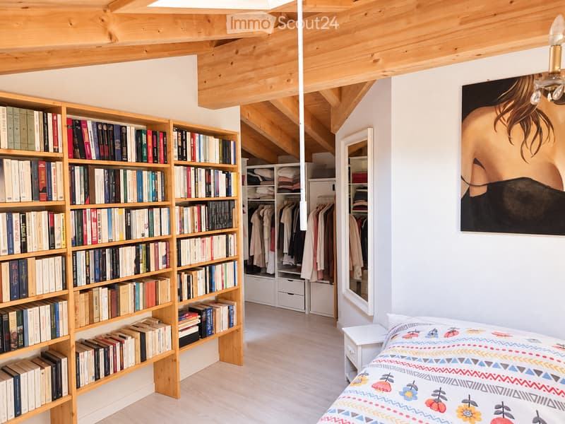 Cozy bedroom with exposed wooden beams, a large bookshelf filled with books, a neatly made bed with a colorful patterned duvet, a small nightstand, a mirror, and a wardrobe with hanging clothes.