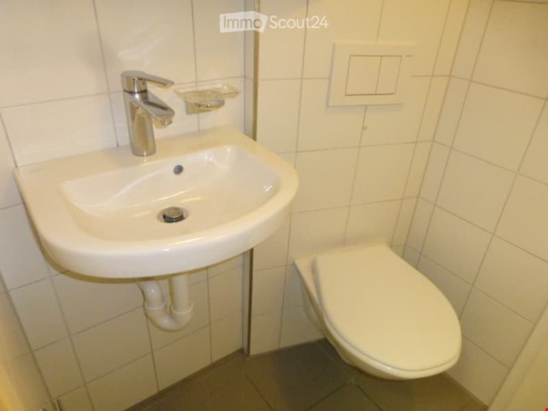 A small, clean bathroom with white tiled walls and floor, featuring a white sink with a modern chrome faucet and a toilet. A soap dish is mounted on the wall above the sink.