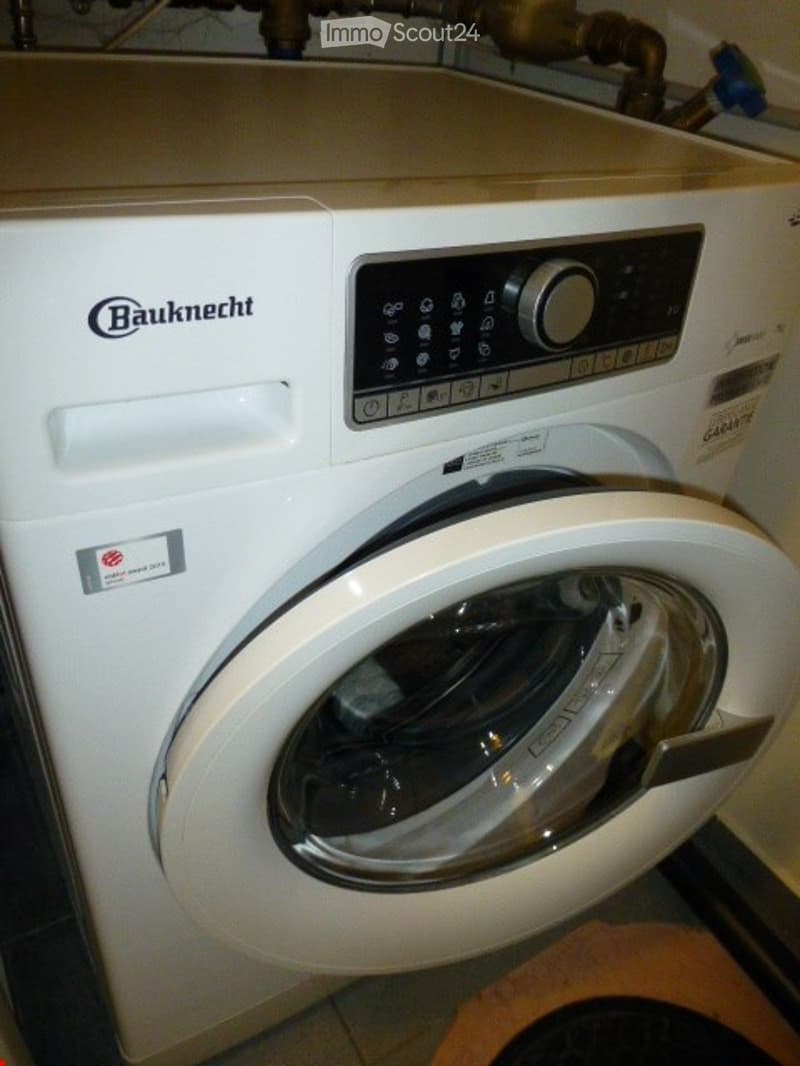 A white washing machine with a control panel and a digital display is installed in a bathroom, with visible plumbing pipes above it.