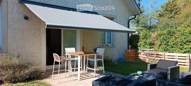 Outdoor patio area with a covered awning, a small table with chairs, and a seating area with a sofa and coffee table, surrounded by greenery and a wooden fence.
