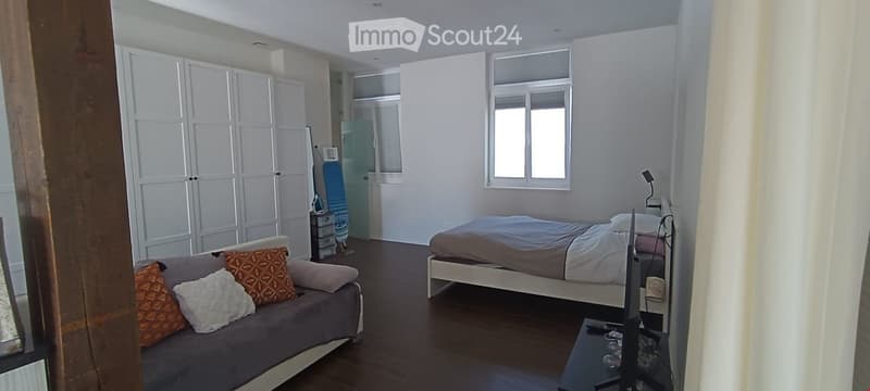A modern, well-lit bedroom with a grey sofa, a bed with a grey duvet, a nightstand, a small table, and a wardrobe with white doors.