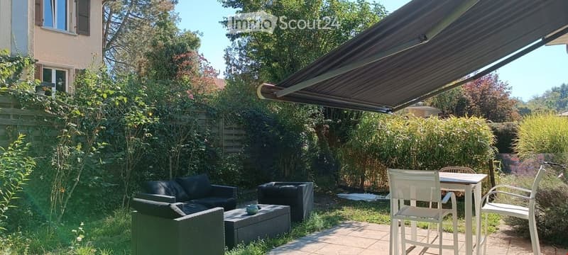 Garden area with outdoor seating, including a sofa, coffee table, and dining table with chairs, surrounded by greenery and a covered patio area.