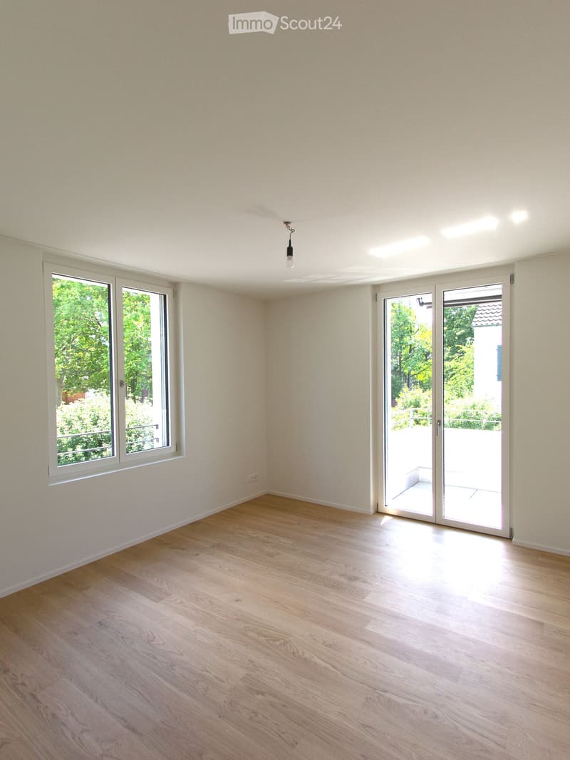 Empty room with light wooden flooring, white walls, large windows, and sliding glass doors leading to a balcony.