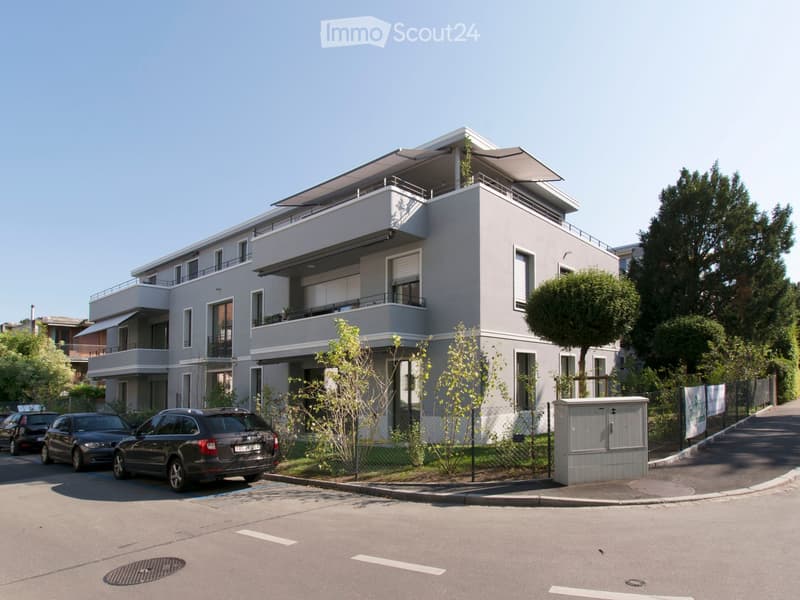 Modern two-story residential building with grey facade, large windows, balconies, and a flat roof terrace. The building is surrounded by greenery and has a parking area with several cars.