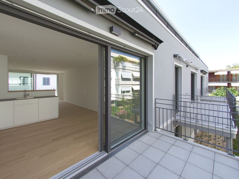 Modern balcony with large glass sliding doors, wooden flooring, white tiled balcony floor, metal railing, and a small kitchen area visible through the doors.