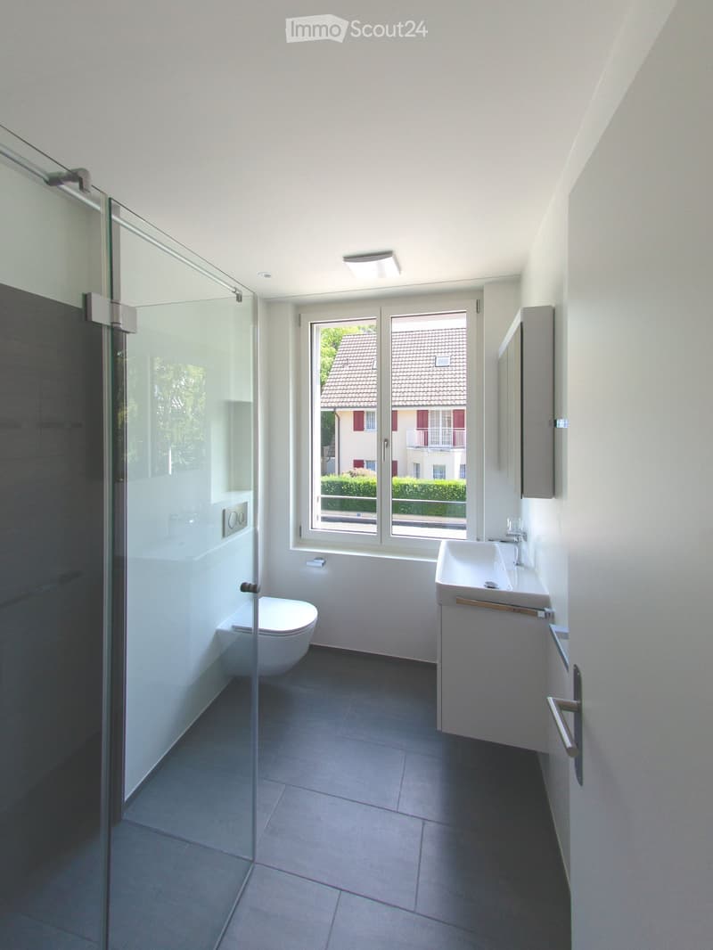 Modern bathroom with a toilet, sink, and glass shower enclosure. The room features white walls, dark tiled flooring, and a large window offering a view of the exterior.