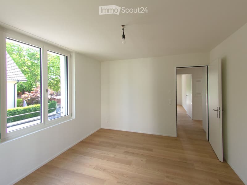 Empty room with light wooden flooring, white walls, large windows, and a pendant light fixture.