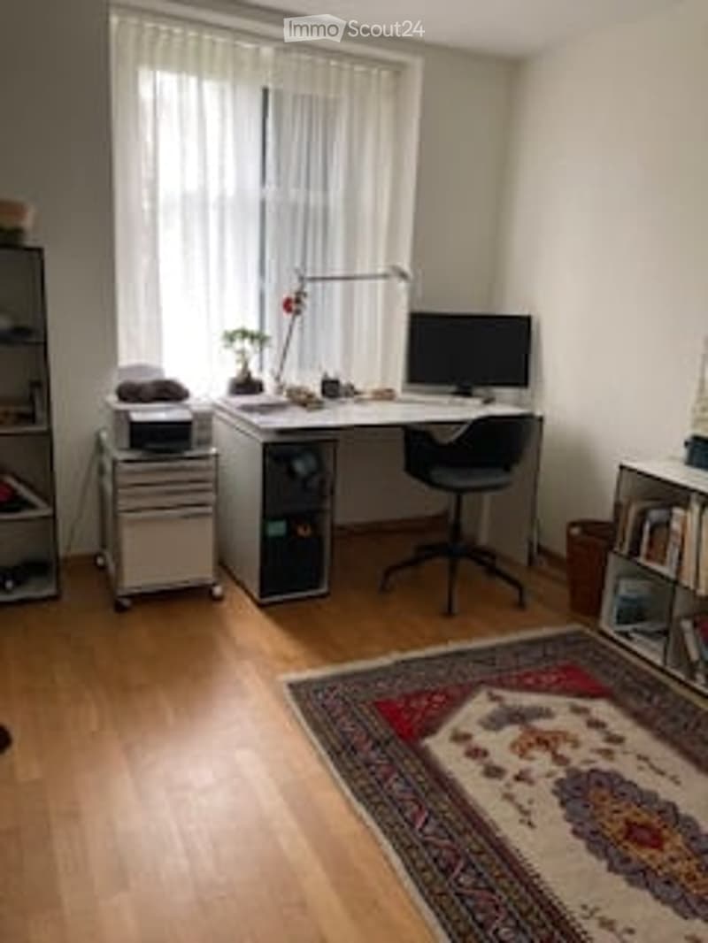 Home office with wooden flooring, large window with white curtains, desk with computer, chair, printer, lamp, small plant, bookshelf, and patterned rug.