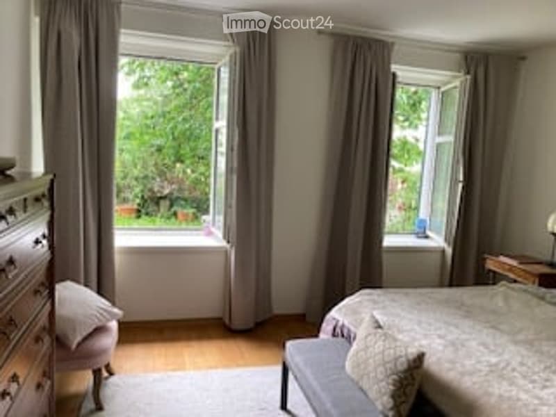 Cozy bedroom with large windows, grey bedding, wooden floor, dresser, nightstand, and a small chair.