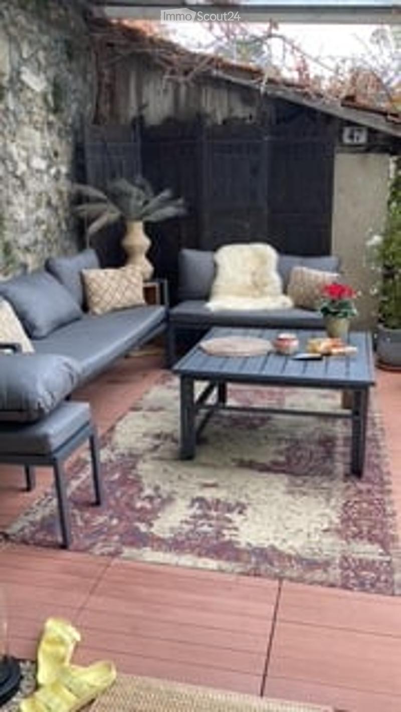 Outdoor patio with a grey sectional sofa, coffee table, patterned rug, and potted plants.