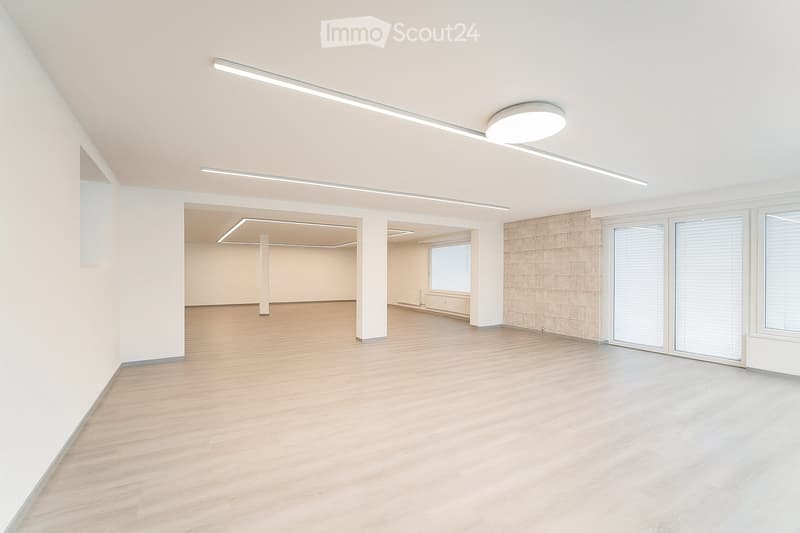 Empty room with light wooden flooring, white walls, and large windows with blinds. The space features modern lighting fixtures and a neutral color palette.
