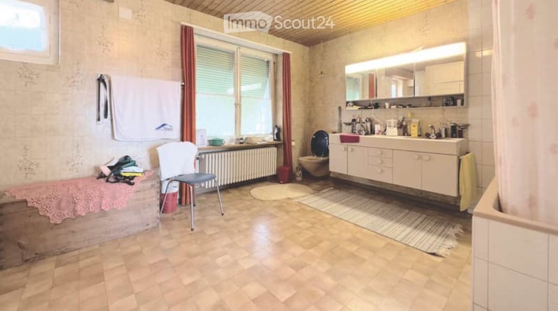 Bathroom with tiled floor, wooden ceiling, large window with red curtains, sink with cabinet, toilet, mirror, chair, radiator, and towels.