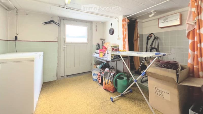 The room is a utility space with a refrigerator on the left, a table with various items including a can, a vacuum cleaner, and an ironing board on the right, and a door leading outside.