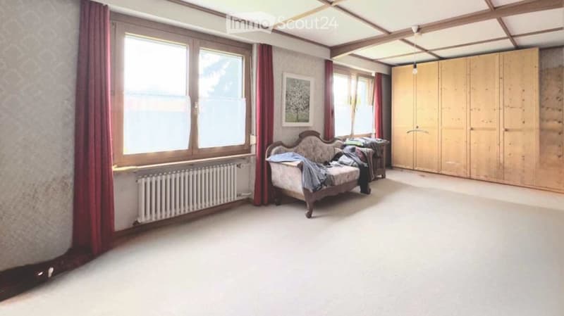 Spacious room with large windows, a patterned sofa, a wooden wardrobe, a small table with items, and a picture frame on the wall.
