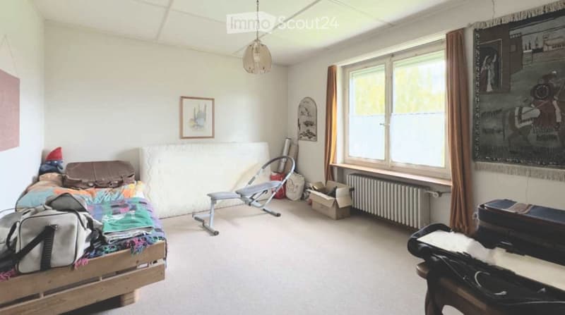 A small, furnished bedroom with a wooden bed, colorful bedding, a chair, a radiator, and a large window with curtains.