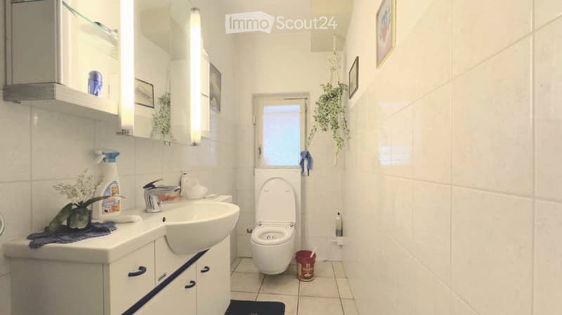 Modern bathroom with white tiles, a sink with a faucet, a toilet, a cabinet, a mirror, shelves, and a small plant on the counter.