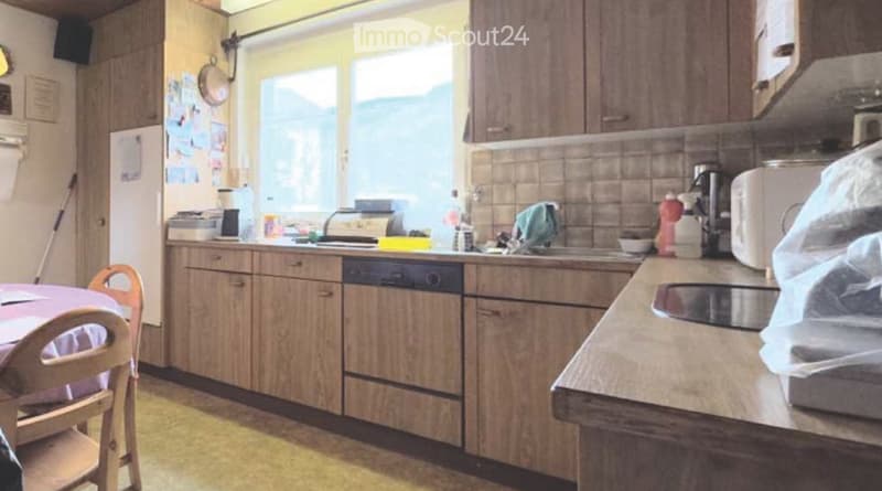 A well-lit kitchen with wooden cabinets, a dishwasher, microwave, and a small dining area with a table and chair. The kitchen features a window, a sink, and various appliances.