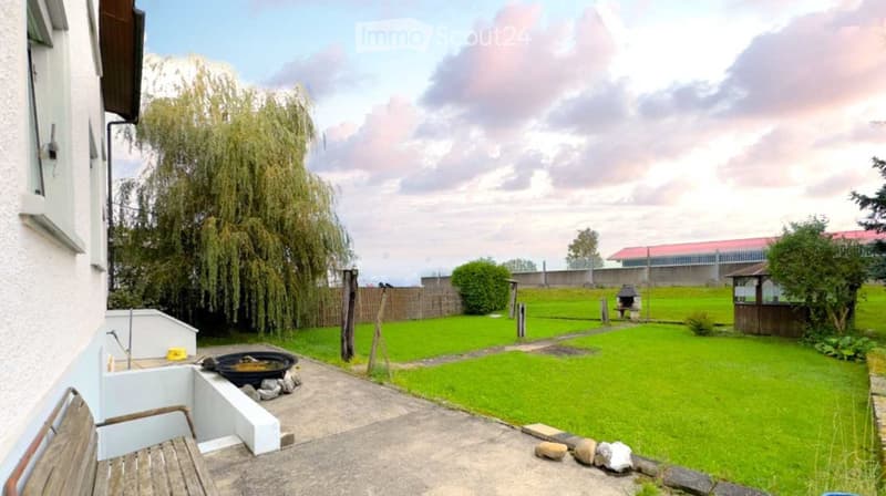 Garden with a well-maintained lawn, a small fountain, a wooden bench, and a barbecue area, surrounded by trees and a fence.