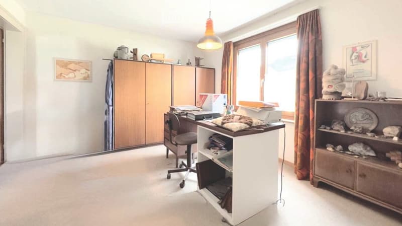 A well-lit room with a desk, chair, cabinet, and shelf, featuring a large window with curtains and a hanging lamp.