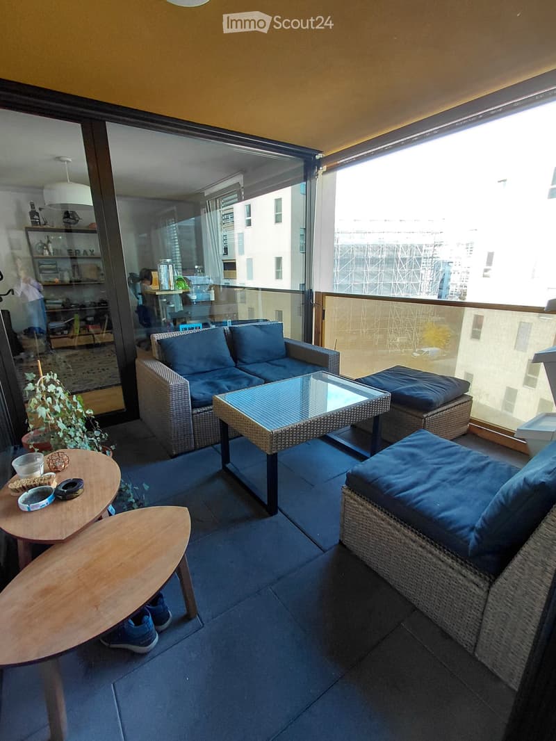 Balcony with a sofa, glass coffee table, wicker chairs, small wooden table, and potted plant.