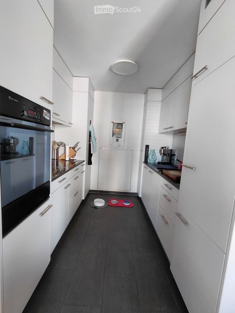 Modern kitchen with white cabinets, stainless steel appliances, and dark tiled flooring. Includes an oven, coffee maker, and various kitchen utensils.