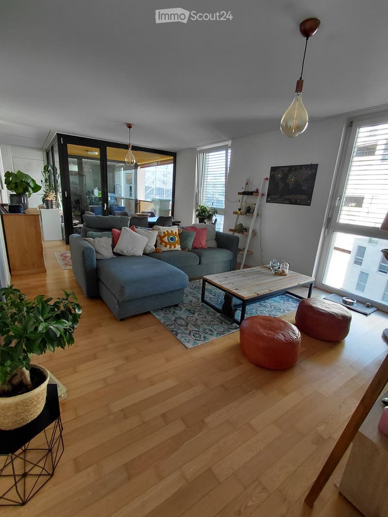Modern living room with light wooden flooring, grey sectional sofa, colorful cushions, coffee table, ottomans, and potted plants.