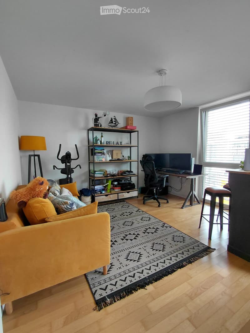 A modern living room with a yellow sofa, patterned rug, wooden floor, desk with laptop, and shelves with books and decorative items.