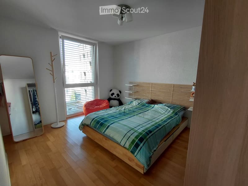 Cozy bedroom with wooden flooring, large window with blinds, bed with plaid duvet, coat rack, mirror, and panda plush toy.
