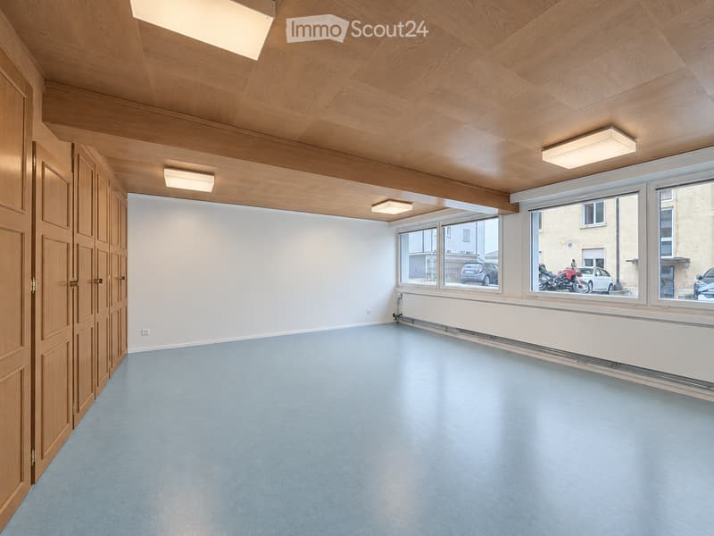 Empty room with light blue carpeted floor, wooden paneled ceiling, white walls, large windows, and a wooden door.