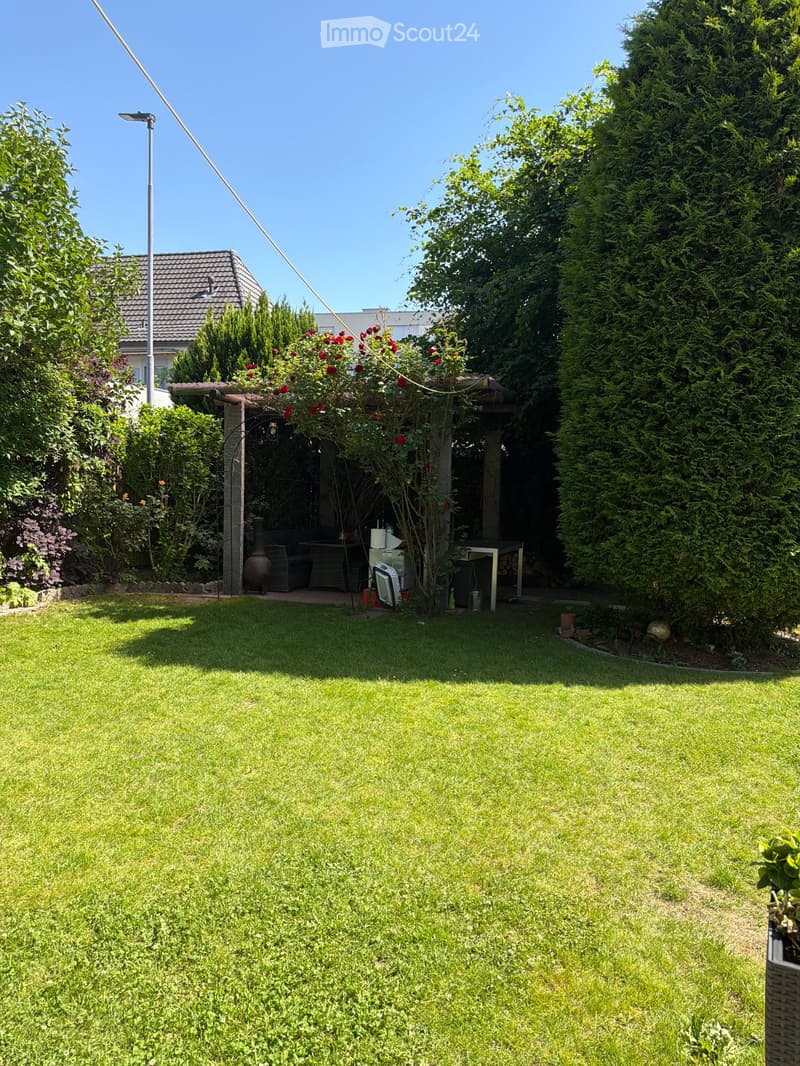 Well-maintained garden with a large lawn, surrounded by trees and shrubs, featuring a patio area with outdoor furniture and a covered seating area.