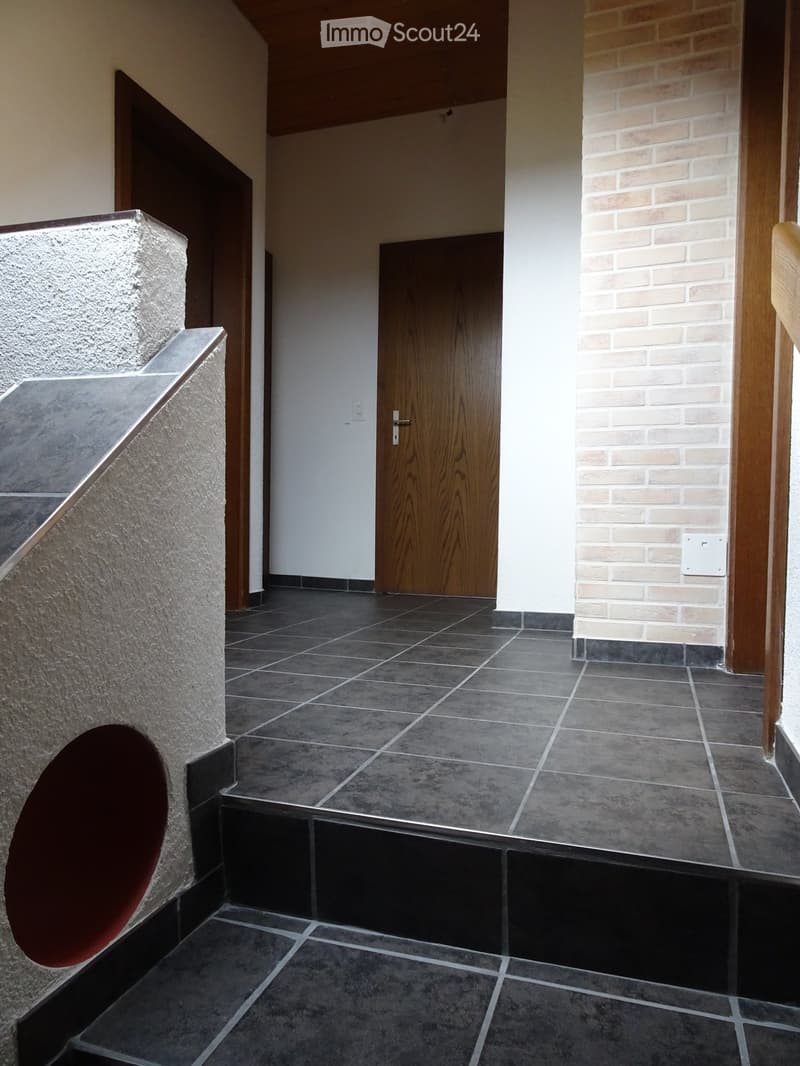 Modern hallway with dark tiled flooring, white walls, and wooden doors. Features a raised section with a curved wall and a brick accent wall on one side.