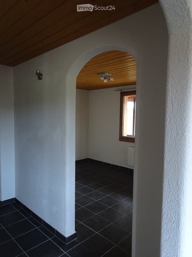 Empty hallway with white textured walls, dark tiled floor, wooden ceiling, and a small window at the end.