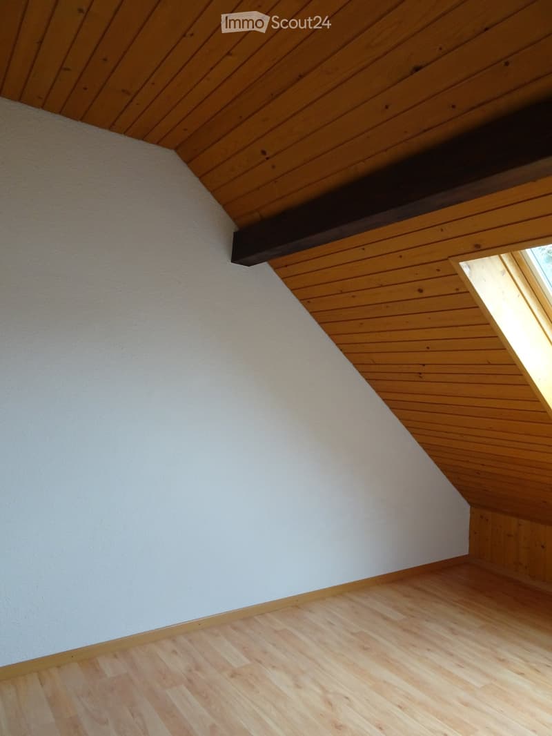 Empty room with wooden flooring and white walls, featuring a slanted wooden ceiling and a small window letting in natural light.