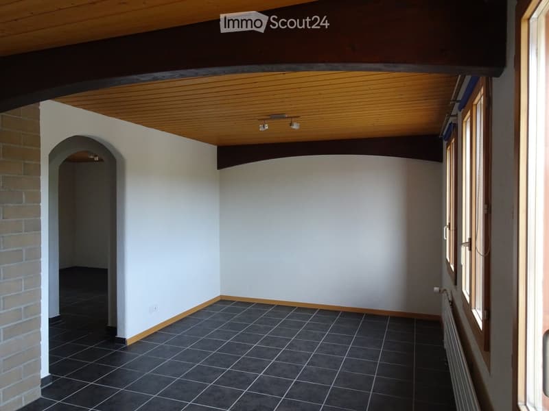Empty room with white walls, dark tiled floor, wooden ceiling beams, and a window with blinds.