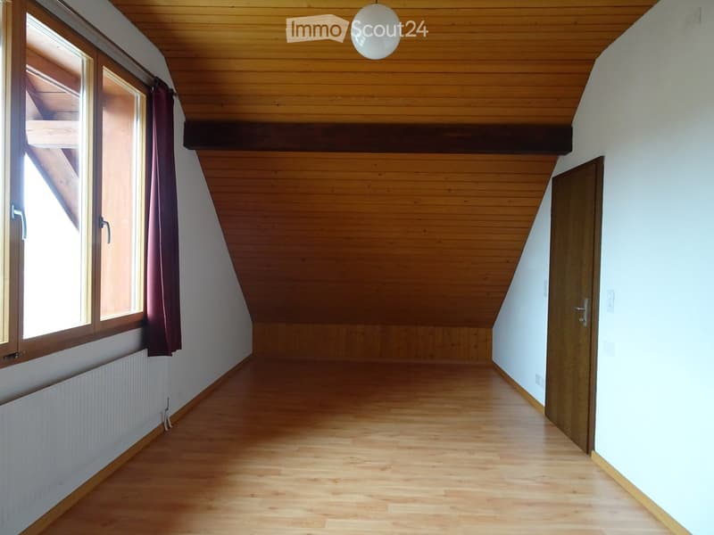 Empty attic room with wooden floors, slanted wooden ceiling, large window with dark curtains, and a closed wooden door.