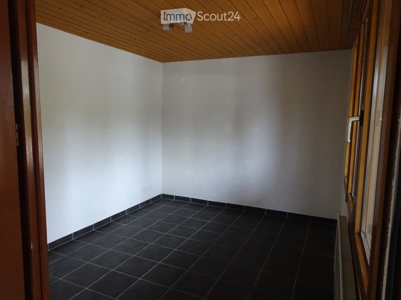 Empty room with white walls, dark tiled floor, wooden ceiling, and a partially open wooden door leading to another space.