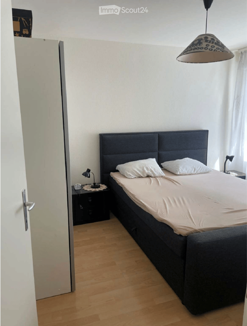 Modern bedroom with a large bed, nightstand, and lamp. The room features light wooden flooring and white walls.