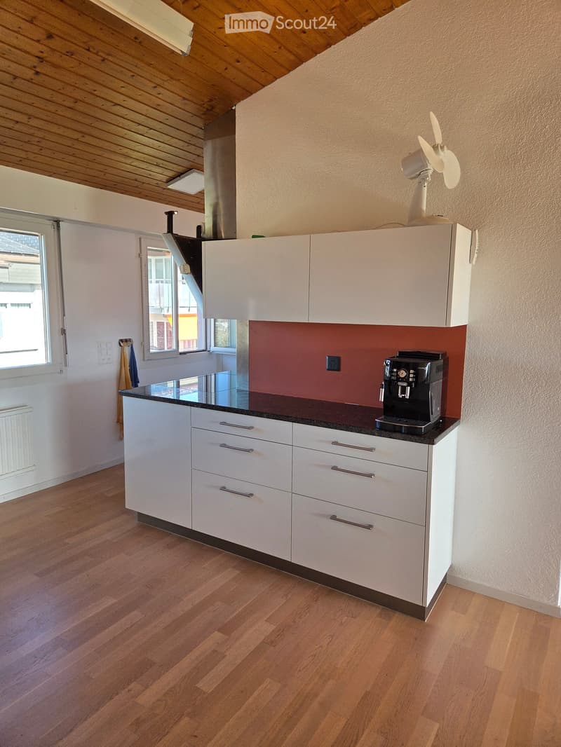 Modern kitchen with white cabinets, black countertop, coffee machine, and a fan on top of the cabinets. The floor is wooden, and there is a window with a view of the outside.