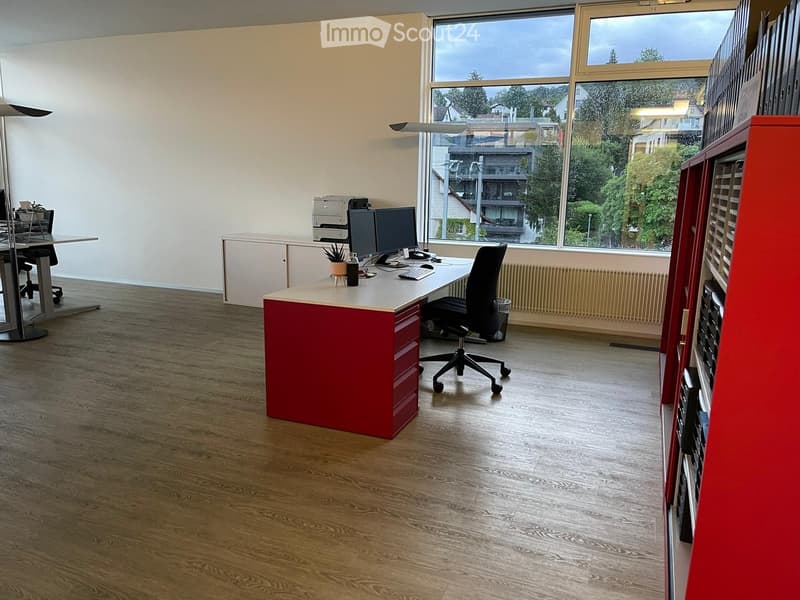 Modern office space with wooden flooring, large windows, a red and white desk, office chair, computer setup, and a cabinet.