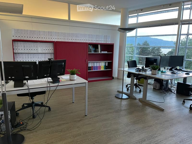 Modern office space with two desks, multiple computer monitors, chairs, a potted plant, and a red cabinet with binders and bookshelves.