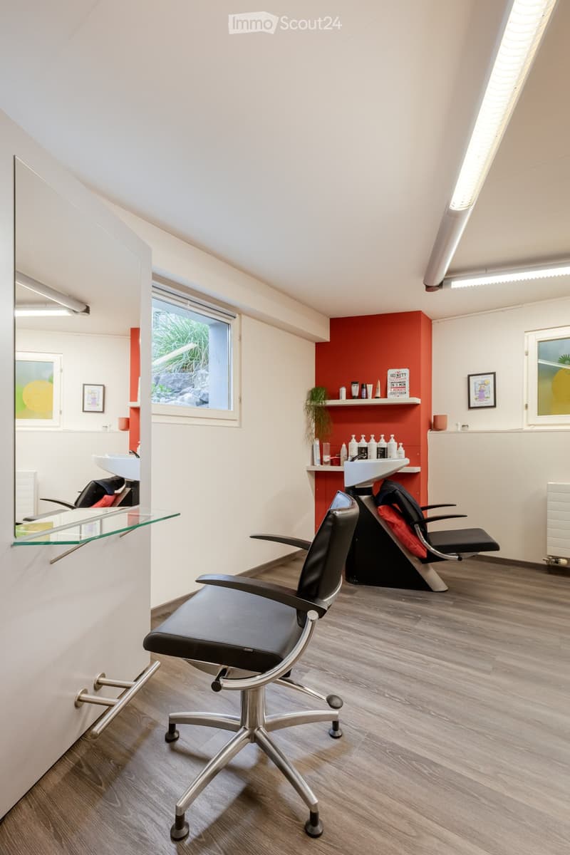 Modern, well-lit room with wooden flooring, two black salon chairs, a sink, shelves with bottles, and framed pictures on the walls.