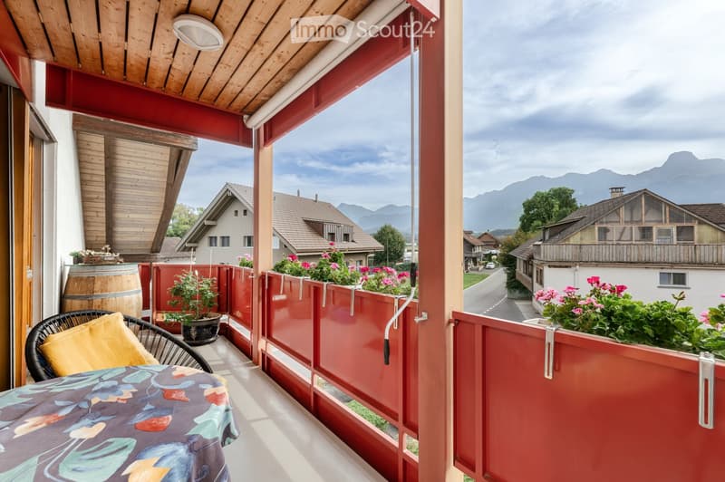 Balcony with wooden ceiling, red railings, table, chair, barrel, and potted plants, overlooking a village with mountains in the background.